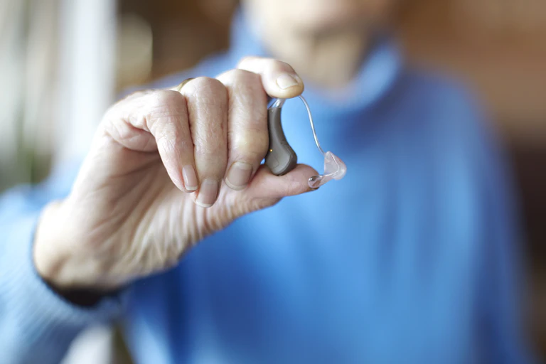 A senior woman, standing in the background, holds up a hearing aid.