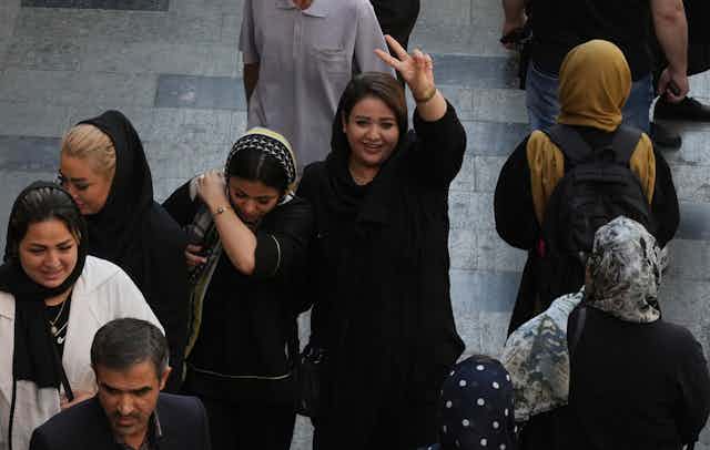 Une femme aux cheveux noirs sourit et fait un signe de victoire en marchant avec d'autres femmes dans un bazar.