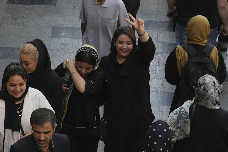 Une femme aux cheveux noirs sourit et fait un signe de victoire en marchant avec d'autres femmes dans un bazar.