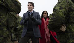 A dark-haired man in a grey plaid jacket stands next to a dark-haired woman in a red coat talking to soldiers in combat gear.