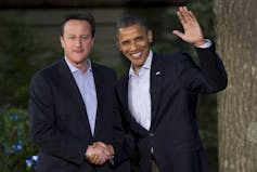 Two men stand together in white shirts and suit jackets. One waves.