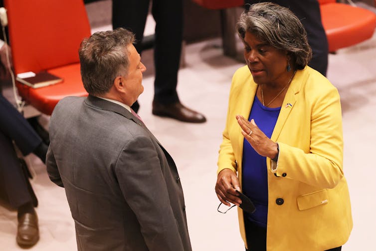 An older white man wearing a gray suit is seen talking to a middle-aged Black woman, who is wearing a yellow jacket and a blue shirt.
