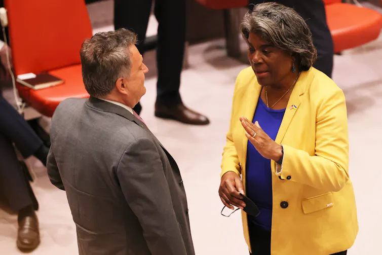 An older white man wearing a gray suit is seen talking to a middle-aged Black woman, who is wearing a yellow jacket and a blue shirt.