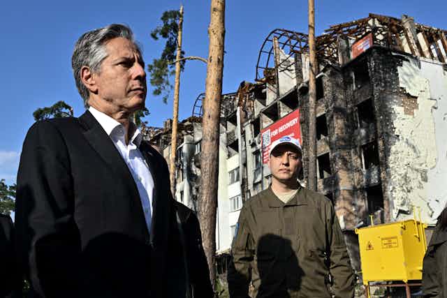 A white middle aged man in a black blazer stands next to a man wearing camouflage shirt, in front of a destroyed, charred building.