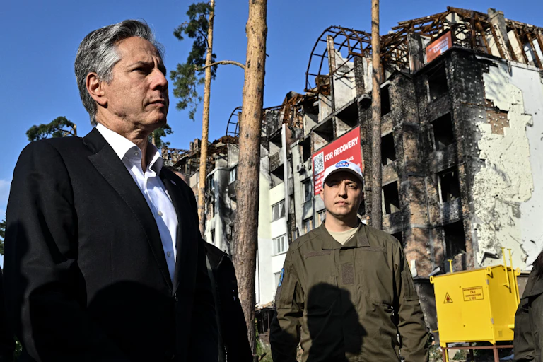 A white middle aged man in a black blazer stands next to a man wearing camouflage shirt, in front of a destroyed, charred building.