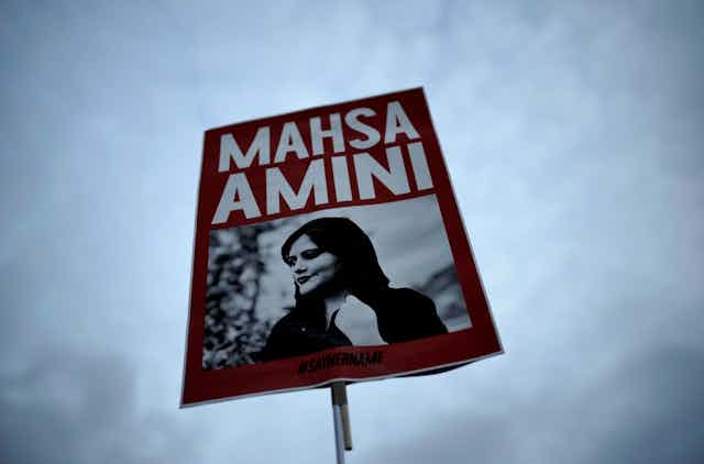 A red placard with a black and white photo of a woman with the words Mahsa Amini.