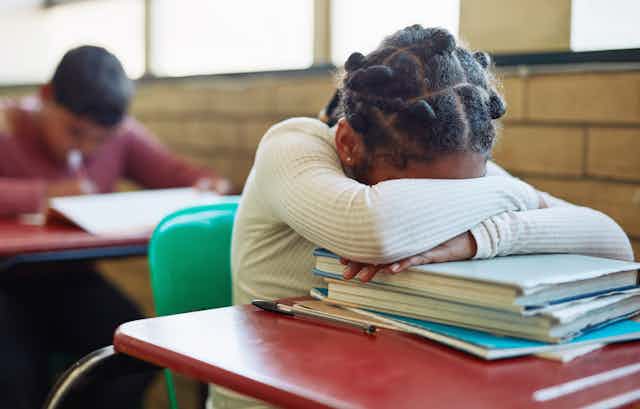 A young female student rests her head on a stack of worn textbooks that sit on the student's desk.