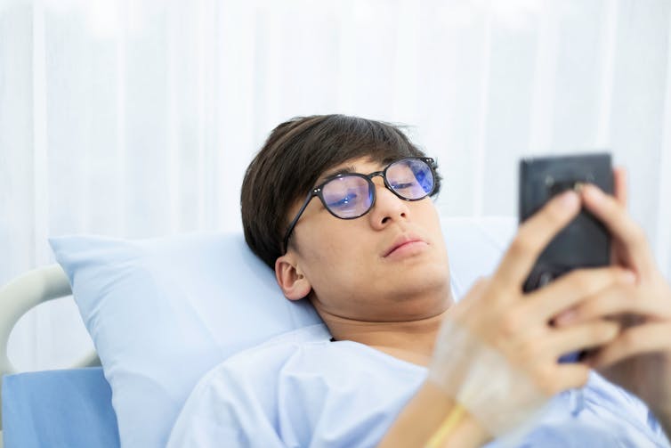 A young man wearing glasses and a hospital gown lying with his head on a blue pillow holding a smartphone with an IV line in his hand.