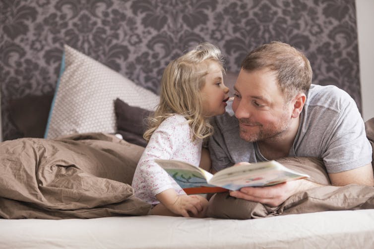 little girl whispers to a man while they read on a bed