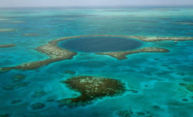 Looking out at the ocean across a region with corals and a large dark blue circle that indicates a blue hole. A large sailboat nearby looks tiny in comparison.