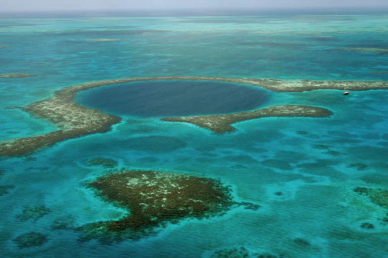 Looking out at the ocean across a region with corals and a large dark blue circle that indicates a blue hole. A large sailboat nearby looks tiny in comparison.