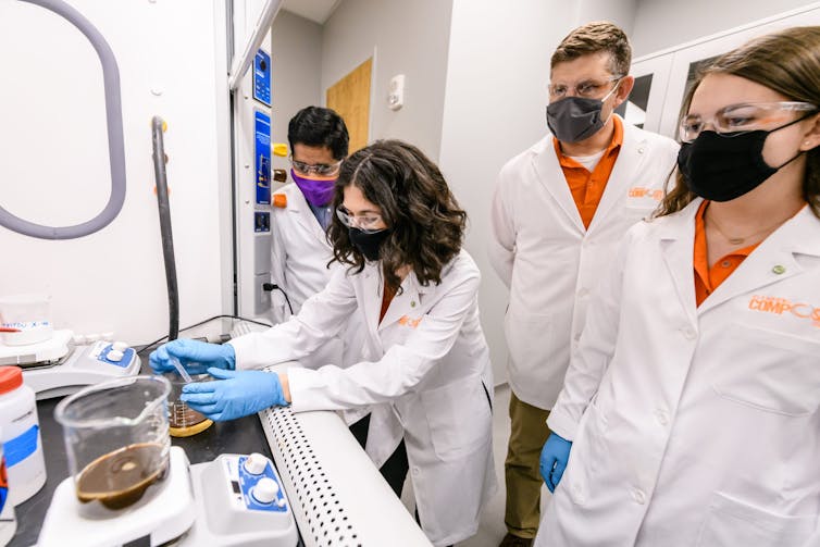 Two men and two women stand over a beaker with dark liquid in it