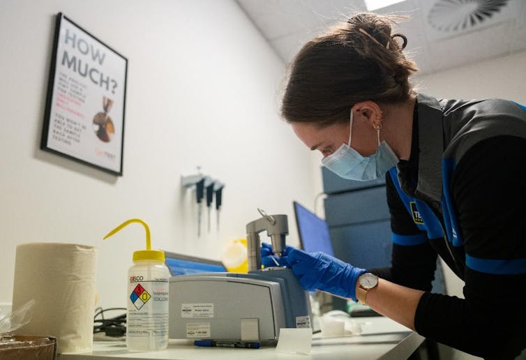 A woman in a lab environment wearing blue gloves leans over a small grey machine