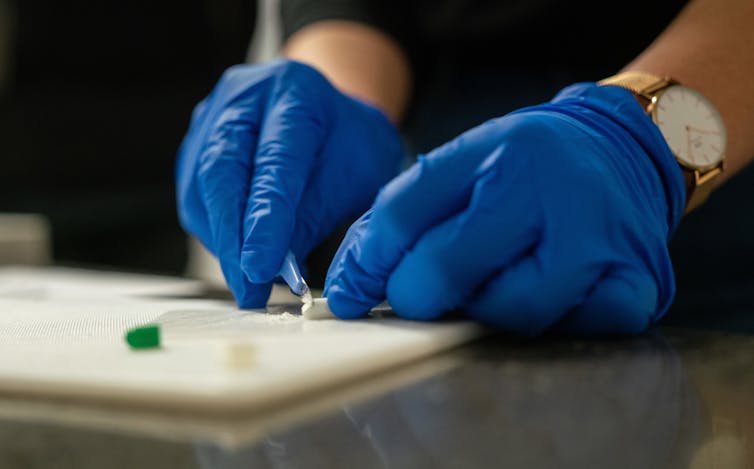 Blue-gloved hands shaving a small amount of white powder off a solid piece