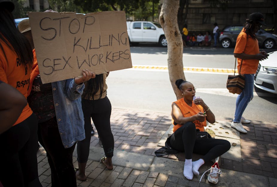 A woman in an orange vest and black pants sits against a tree in her socked feet. shoes at her side. Beside her, a person holds up a sign reading "Stop killing sex workers". Their face is obscured by the sign