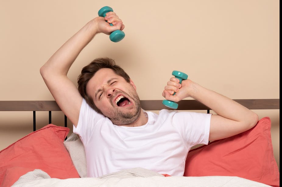 Man holding two dumbbells while lying in bed yawns.