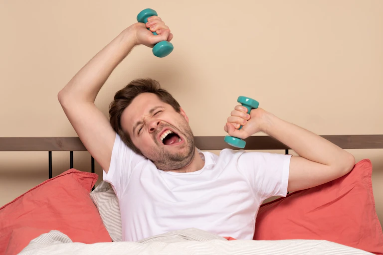 Man holding two dumbbells while lying in bed yawns.
