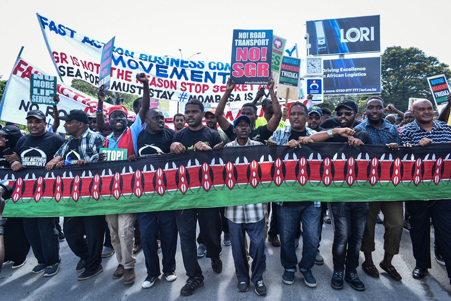 A crowd of men walking on a road while holding the Kenyan flag and placards written 'No SGR', 'No More Lip Service'.