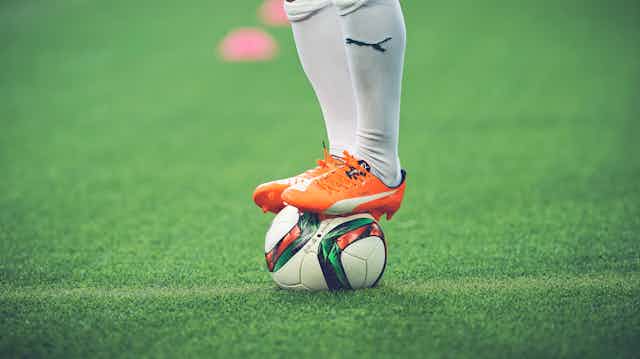 A football player's legs wearing white socks and orange shoes with the Puma logo standing on top of a football.