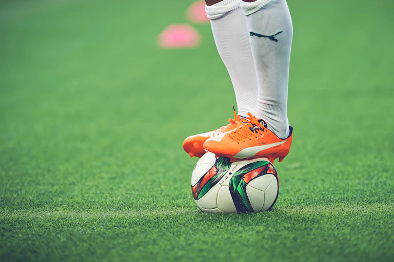 A football player's legs wearing white socks and orange shoes with the Puma logo standing on top of a football.