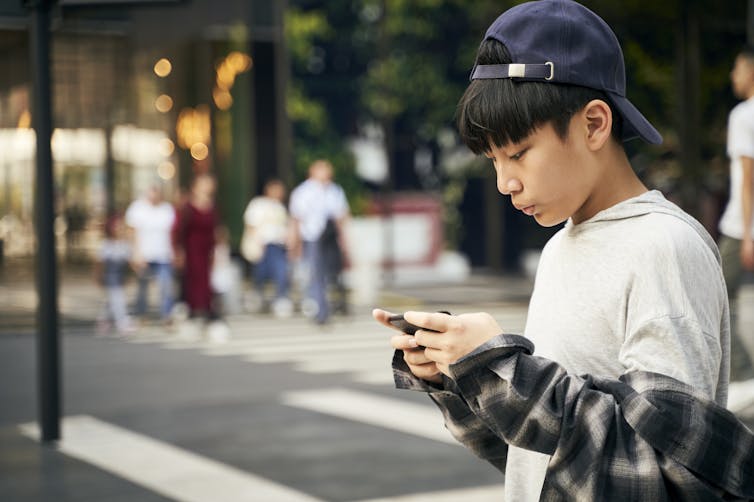 Boy in baseball cap looking at his phone outside on street corner.