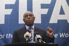 A man wearing a suit stand at a podium. Behind him is a large sign that says FIFA.