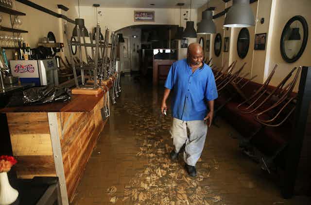 A man walks through a muddy restaurant that had been flooded. Stools are stacked on the counter.