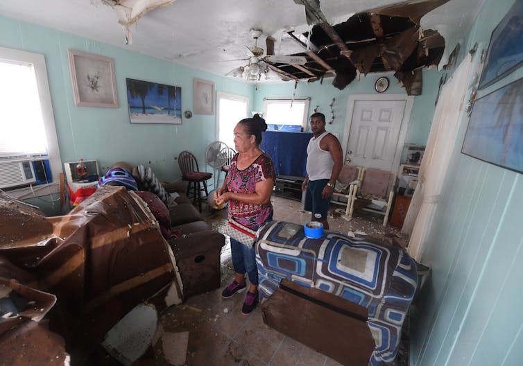 An Hispanic woman and man stand in a flood-damaged living room. The ceiling has a hole from the storm.