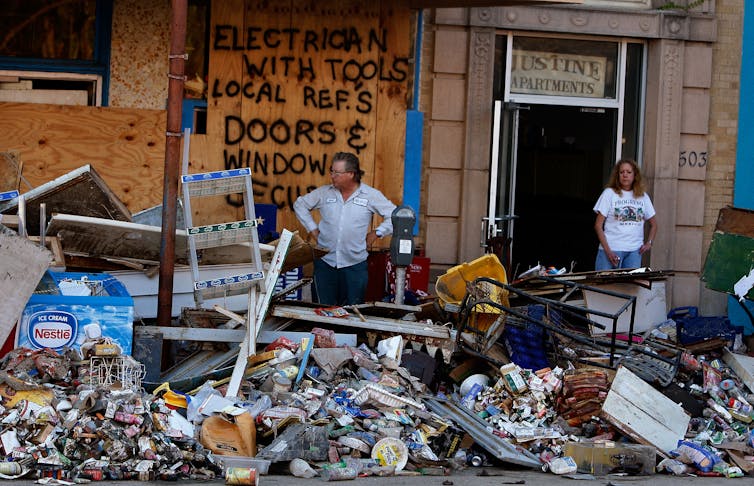 A man stands with his hands on his hips looking a wet household goods that have been dragged out into the street in front of an apartment building.