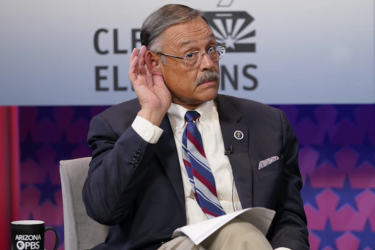 A man in a white shirt, blue blazer and red, white and blue tie cups his ear to hear something while sitting down.