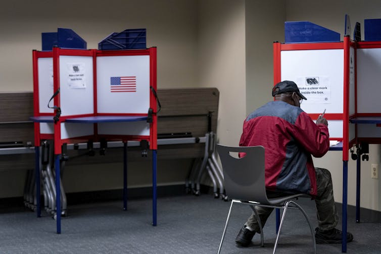 A man in a gray and red jacket wearing a baseball cap sits down at a voting station to cast his ballot.