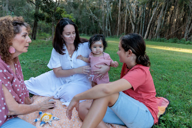 A First Nations family sit together outside. A woman holds a baby. They wear ochre on their faces.