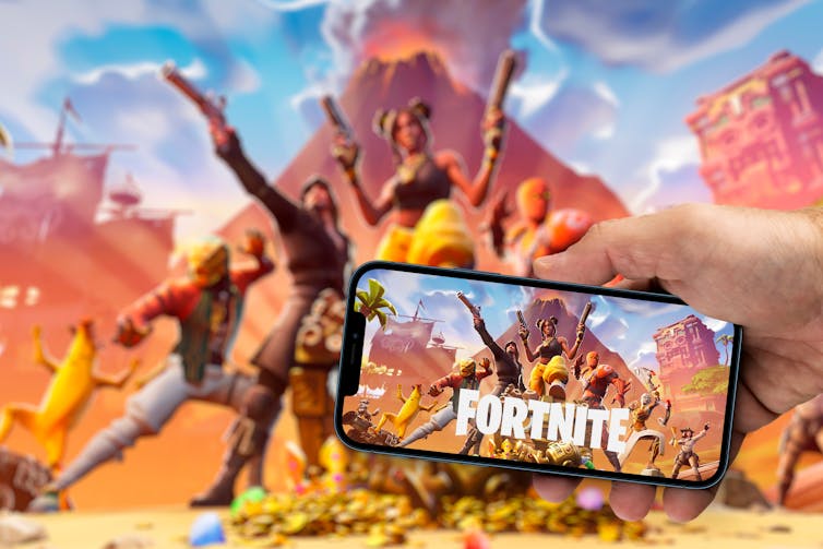 A mobile phone is help up against a background screen. Both show a Fortnite video game title screen.