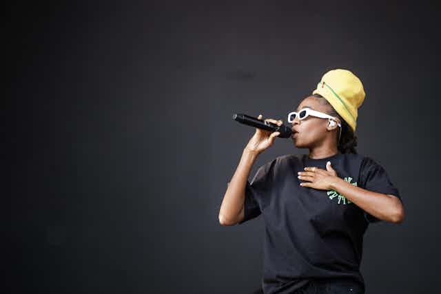 Young black rapper Little Simz singing on stage in sunglasses and yellow hat.