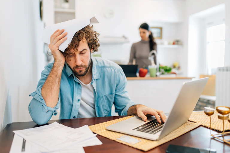 A man calculating bills at a laptop in a kitchen with a woman in the background.