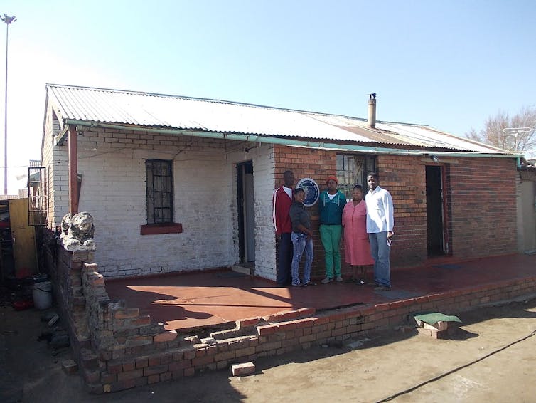 A modest old brick house with a tin roof. A group of people stand having their photo taken at its heritage plaque.