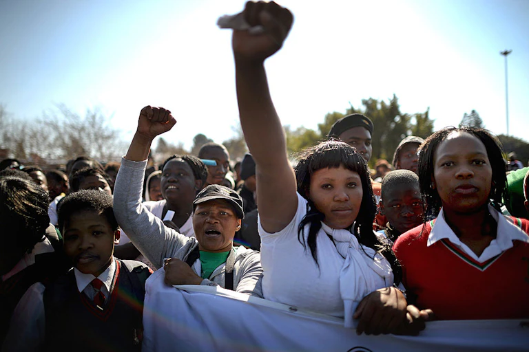 A crowd of people holding a banner, in the front a young woman raises her fist in the air.