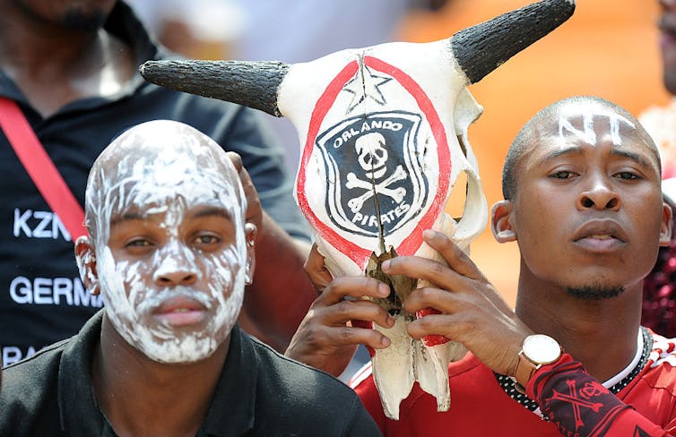 Two men hold a bull's skull painted with the skull-and-crossbones logo of Orlando Pirates football club. They have white face paint on.