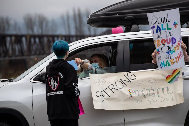 A teen with blue hair seen facing a car with a rainbow poster.