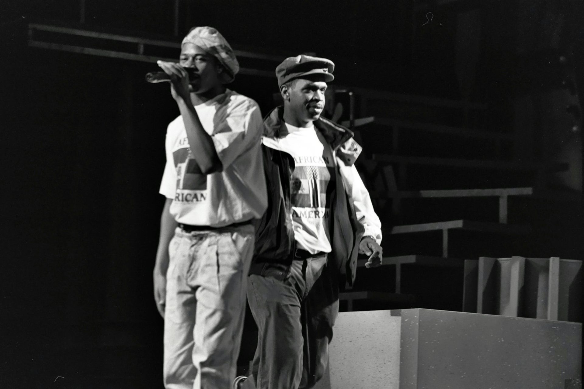 Two young Black men wearing hats stand back to back on a stage.