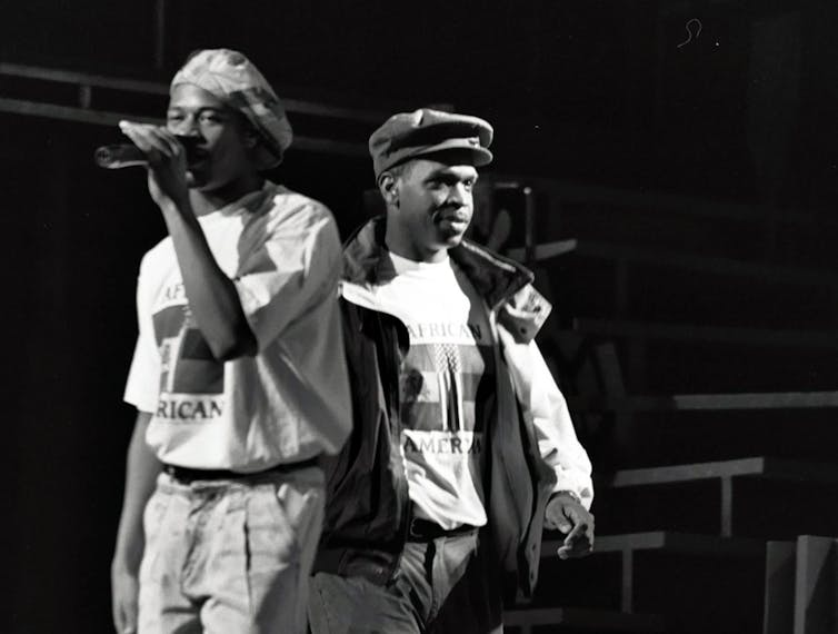 Two young Black men wearing hats stand back to back on a stage.