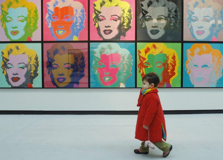 A toddler in a red coat walks by a display of colorful variations of a Marilyn Monroe portrait
