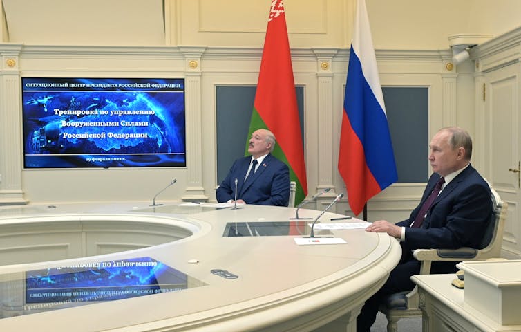 Vladimir Putin and Alexander Lukashenko sit at a round table watching a video screen.