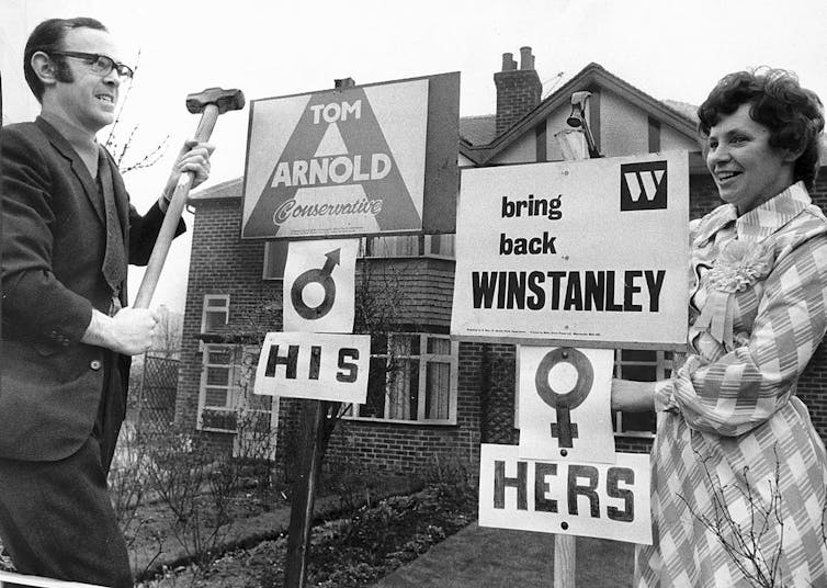 A husband and wife pose next to signs on poles representing competing candidates.