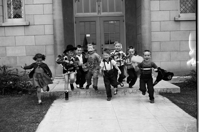 black and white photo of schoolchildren in halloween costumes running out the front door of school
