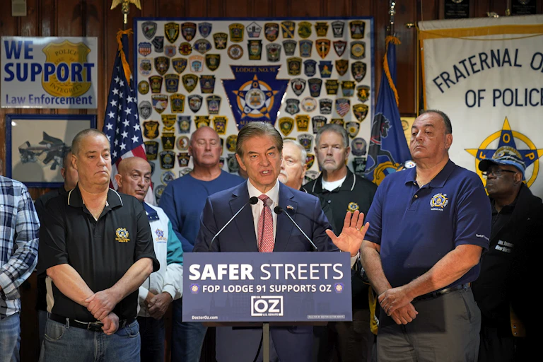 A man stands in front of a lectern with two microphones, flanked by men in short sleeve shirts
