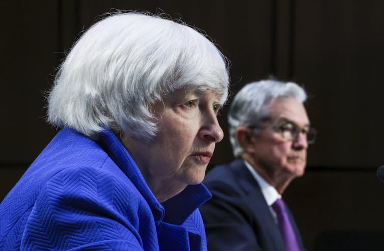 A white woman with gray hair and a blue shirt is seen next to a white man with gray hair, glasses and a dark suit and purple tie