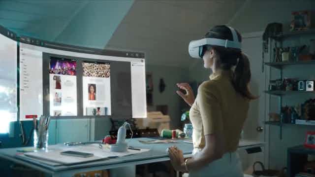 A photo of a woman wearing a VR headset looking at a virtual computer monitor floating above a desk.