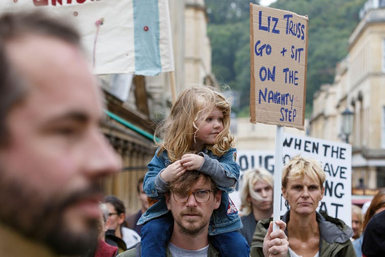 A little girl sits on a man's shoulders in a crowd of protestors holding banners.