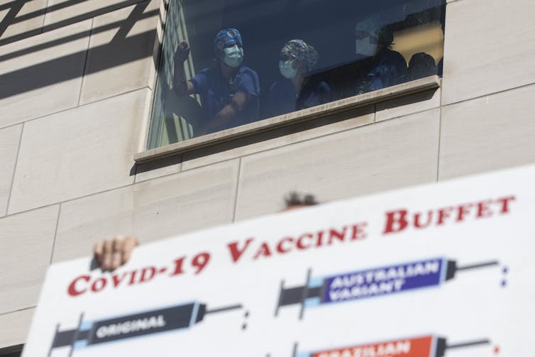Health-care workers in scrubs and masks look out a window at the top of protest signs.